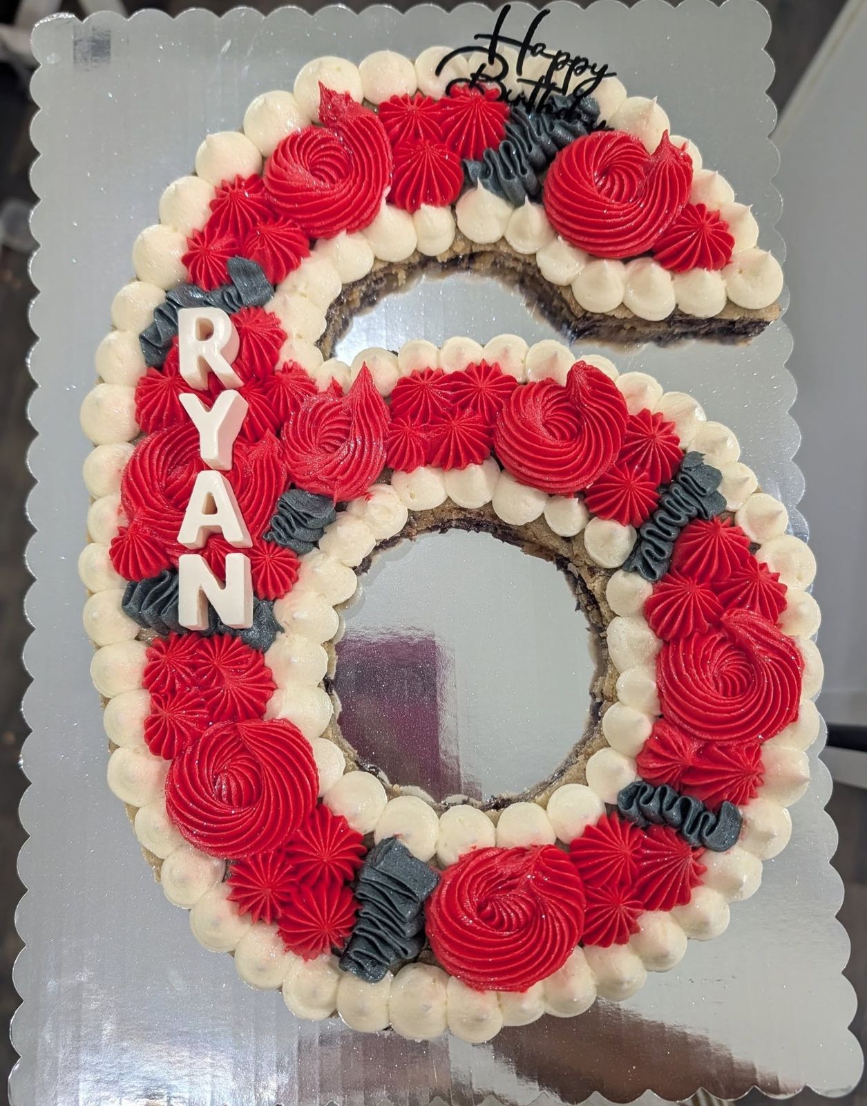 Number 6 cookie cake with red rosettes, cream and navy piping and the name RYAN